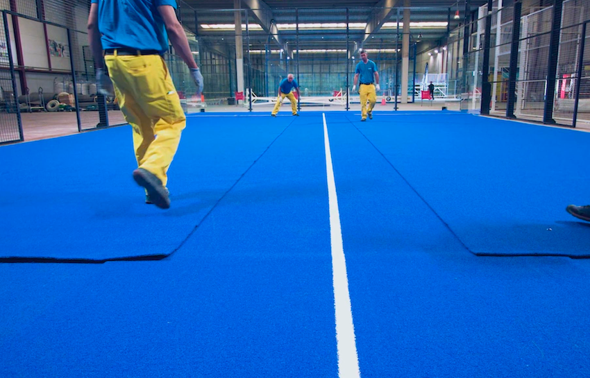Two workers installing blue artificial turf for an indoor padel court.