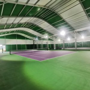 Interior view of a modern clear span aluminum sports tent used as an indoor tennis court, featuring professional LED lighting and a curved roof design.